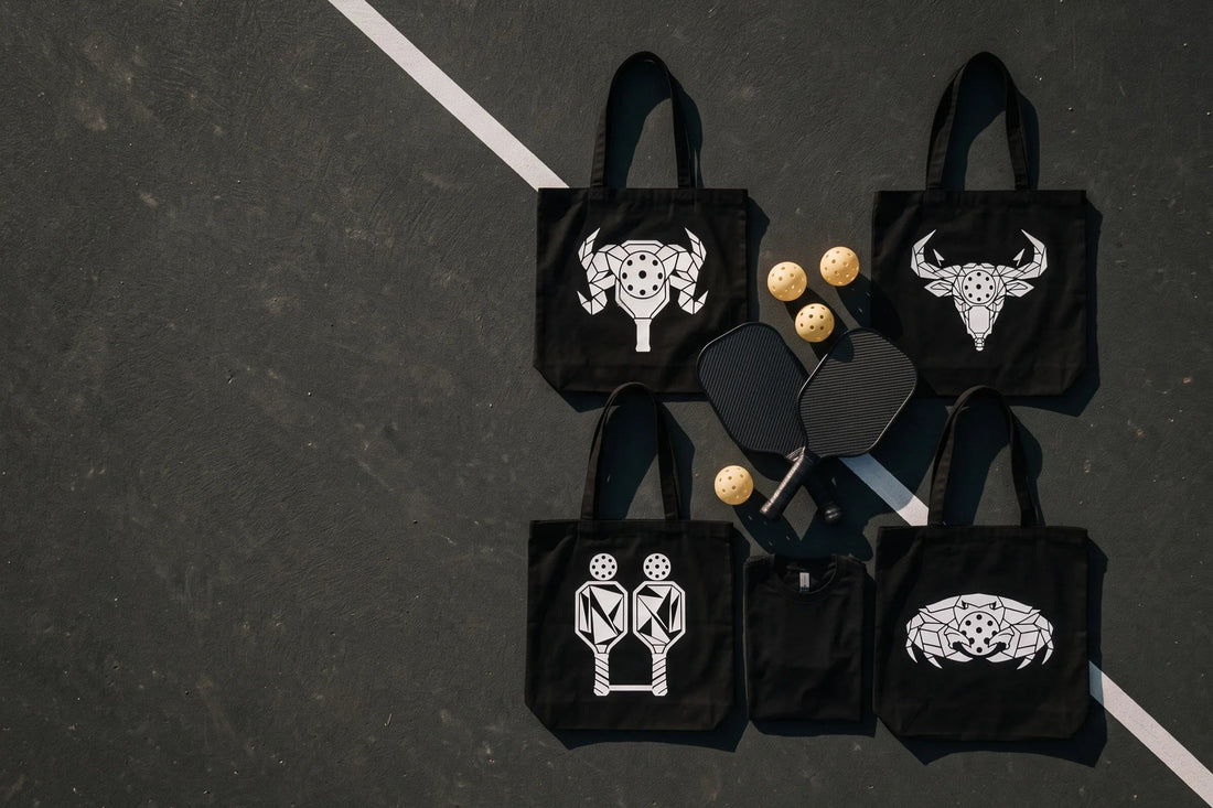 Four black zodiac pickleball tote bags from LMMM Studio Sign Language Drop 02 laid on a dark court with paddles and balls